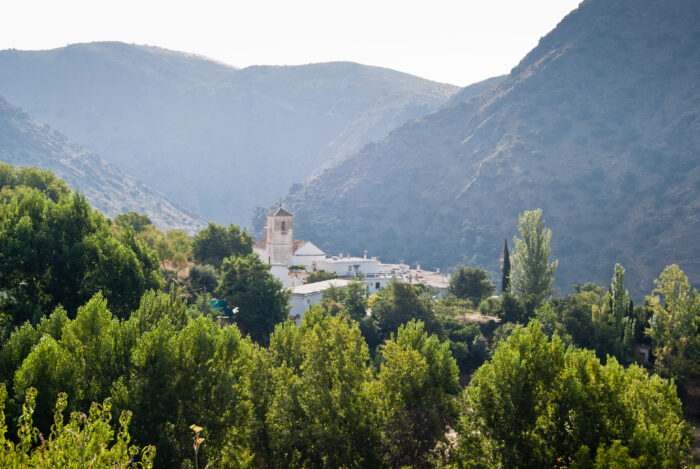 The small village of Ferreirola, with white buildings and a church tower, is nestled among green trees, surrounded by tall, misty mountains under a bright sky.