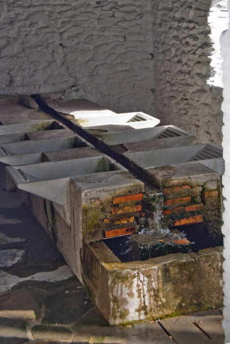 A rustic stone and brick communal wash-house in Ferreirola with water flowing through channels, set in a whitewashed, stone-walled room.