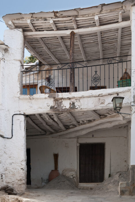 A rustic whitewashed building in ATALB&Eacute;ITAR with a sloping wooden roof, an upper balcony with black wrought-iron railing, and a lower entrance partially surrounded by stone and clay pots.