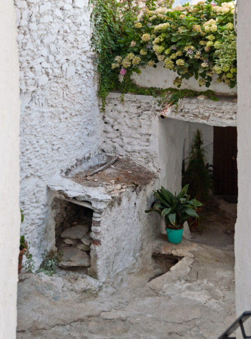 A narrow stone alleyway in ATALB&Eacute;ITAR with whitewashed walls, potted plants, and overhanging greenery. The space appears rustic and aged, with a weathered structure and flowering plants above.