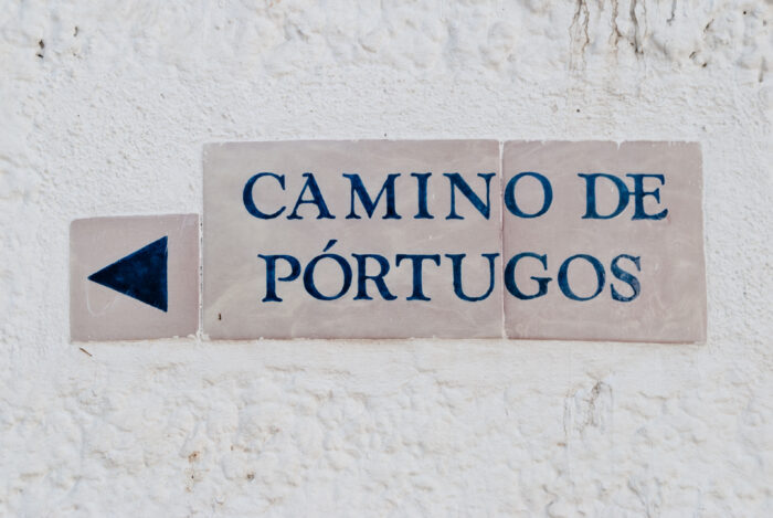 A ceramic sign on a white textured wall reads &ldquo;CAMINO DE P&Oacute;RTUGOS&rdquo; in blue letters with a left-pointing arrow on the left side.