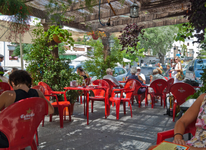 People sit at red plastic chairs and tables under a shaded patio at an outdoor caf&eacute; in Pitres. Lush green plants hang and grow around the seating area, and sunlight filters through the overhead covering. Cars and trees are visible outside.