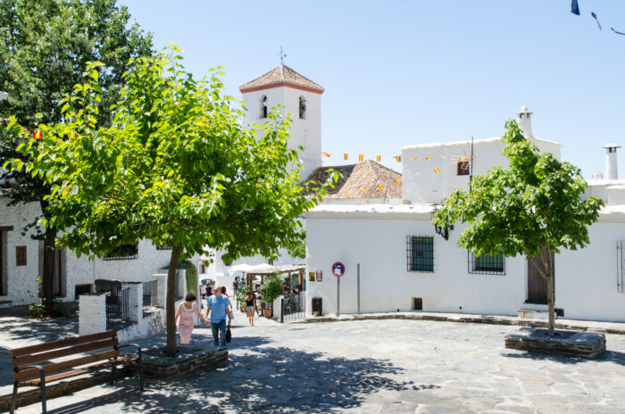 A sunny village square in Pitres with white buildings, leafy green trees, and a stone pavement. People walk towards a small church with a bell tower, and colourful flags hang above. A wooden bench sits in the foreground.