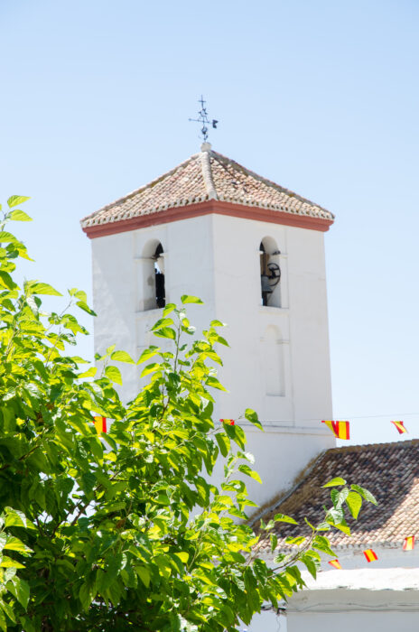 A white bell tower in Pitres with a tiled roof and weathercock rises behind green foliage, with small red and yellow flags strung across in the foreground under a clear blue sky.