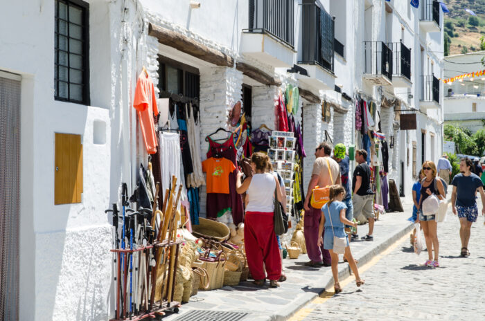 People browse colourful clothes, walking sticks, and souvenirs displayed outside whitewashed shops on a sunny, cobbled street in Capileira.