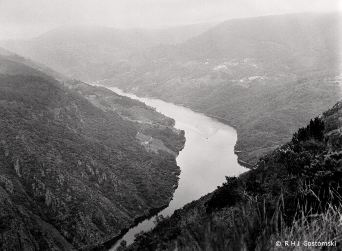 Black and white photo of the Ca&ntilde;&oacute;n del Sil in Galicia, with a winding river cutting through steep, forested hills and valleys, misty mountains in the background, and scattered buildings dotting the slopes.