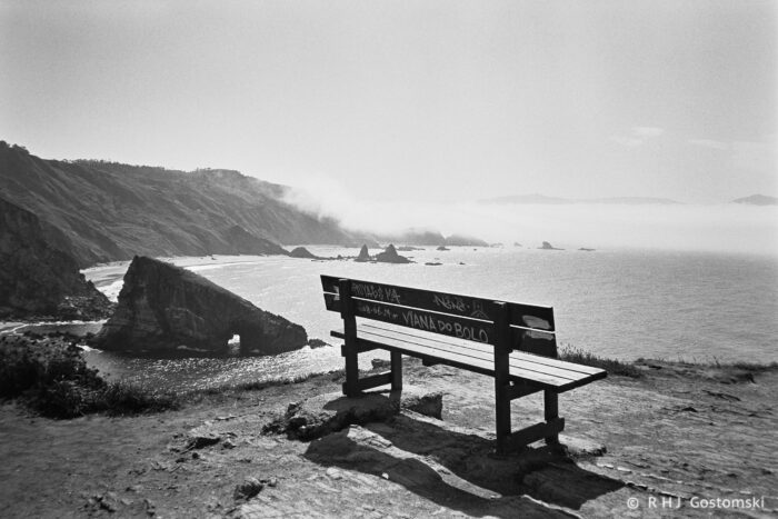 A wooden bench with graffiti overlooks the rugged Loiba coastline of Galicia, where cliffs and sea meet. Mist rises over the distant shore, and sunlight creates a hazy, tranquil atmosphere.