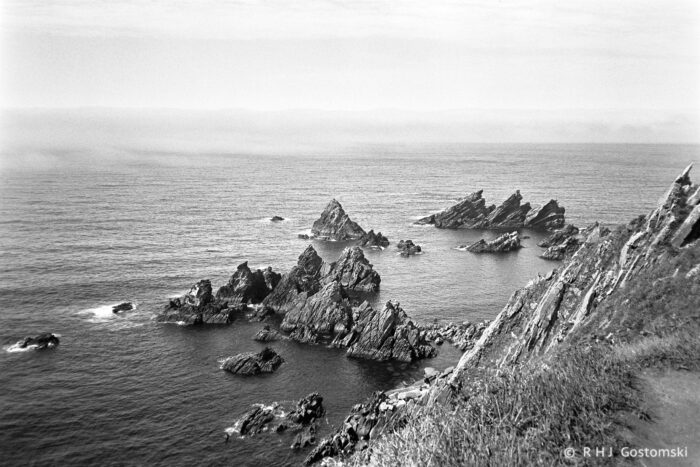 Black and white photo of jagged rocks jutting out of the sea near a steep cliff along the wild Loiba coastline in Galicia, with gentle waves and a hazy horizon in the distance.