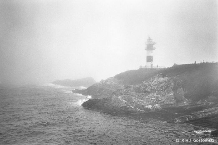 A foggy coastal scene near Ribadeo, with waves crashing against rocky cliffs and the Illa Pancha lighthouse standing on the headland. The image has a misty, atmospheric quality, softening the landscape and obscuring distant details.