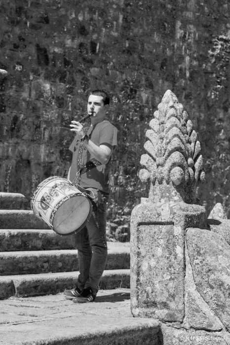 A man playing a traditional flute and carrying a drum stands on stone steps leading to the church A man playing a traditional flute and carrying a drum stands on stone steps next to an ornate stone sculpture, with a textured stone wall in the background. The image is in black and white.