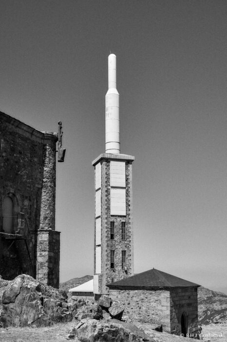Telecommunications repeater antenna A tall stone tower with a white cylindrical telecommunications repeater aerial stands beside an old stone building, set against a clear sky and rocky hillside in a striking black and white landscape.