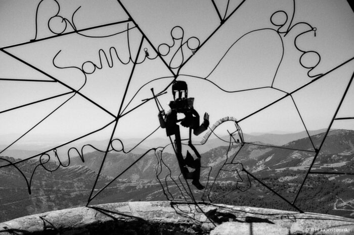 View from the Balcón de Santiago A wire sculpture of a Santiago pilgrim holding a staff on a wire horse sculpture, with handwritten-style wire words and distant mountains visible in the background under a clear sky.
