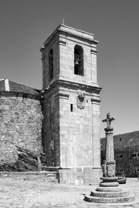 The Peña de Francia square and the bell tower dating back to 17 Black and white photo of an old stone bell tower next to a stone wall. In the foreground, there is an ornate stone cross on a pedestal set in a cobbled plaza under a clear sky.