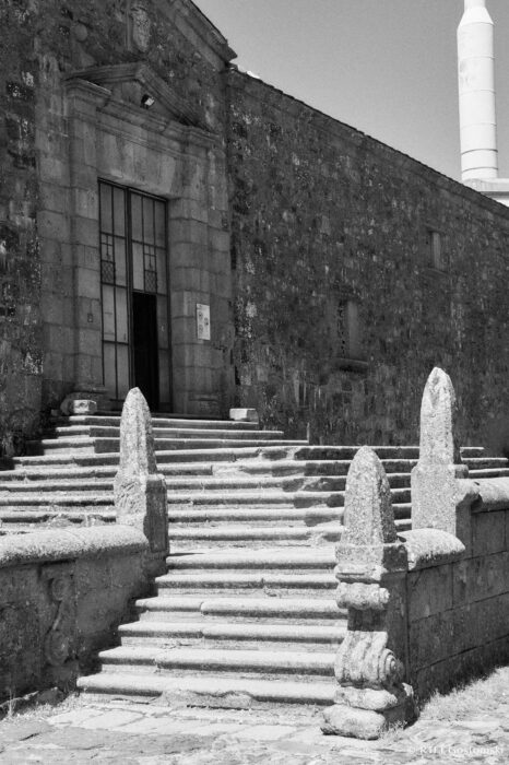 Entrance to the church of the Virgin of the Peña de Francia A black and white photo of the church of the Virgin of the Peña de Francia with a large door and a set of wide stone steps leading up, flanked by carved stone posts. A tall telecom transmitter tower is visible in the background on the right.