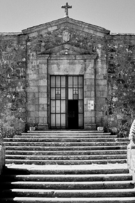 Entrance to the church of the Virgin of the Peña de Francia Black and white photo of the entrance to the church of the Virgin of the Peña de Francia. Symmetrical steps lead up to the large double entrance doors. The stone facade shows weathering and age.