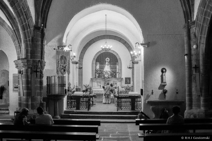 Inside the Virgin of the Peña de Francia church Black and white photo of a church interior with arched ceilings, a central altar featuring a statue of Mary, people sitting on pews, and a couple standing near the altar, softly lit by chandeliers and wall lamps.