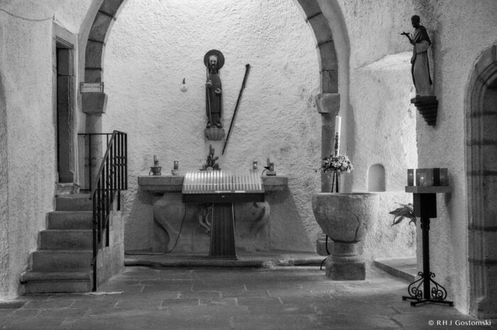 The Santiago chapel. Black and white photo of the Santiago chapel altar at Peña de Francia, featuring a saint’s statue on the wall, stone arch, small staircase, baptismal font, flowers, and another standing figure statue to the right.