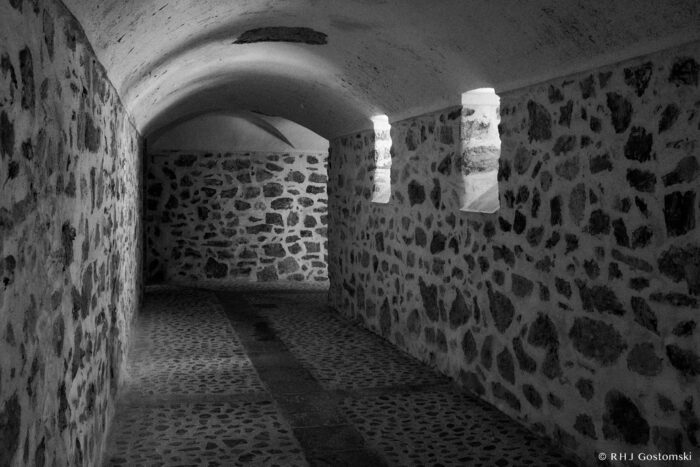 Access to the Pozo Verde (green well) A black-and-white photo of a narrow, arched stone corridor with cobblestone floor, leading to the Pozo Verde (green well). Two wall-mounted lights on the right side illuminate the textured walls, creating shadows and a slightly eerie atmosphere.