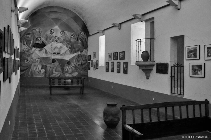 A room in the convent Black and white photo of an romm in the convent with paintings on the walls, a mural of the Last Supper at the far end, benches, an amphora on the floor, and a small balcony with a vase on the right-hand side.