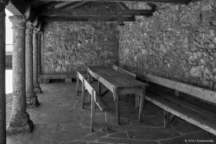 Columns and a sheltered area by Rollo Square Black and white photo of a rustic stone-walled room with a long wooden table, benches on both sides, stone columns to the left, and a rough stone floor.