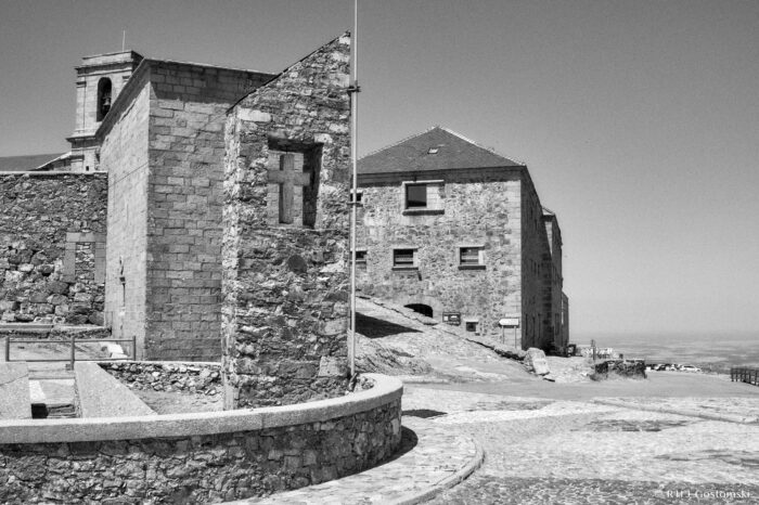 The Rotunda, also known as the Mirador de Santo Domingo Black and white photo of a historic stone building and cross monument on a cobbled Mirador de Santo Domingo on Peña de Francia, with distant views of the landscape under a clear sky.