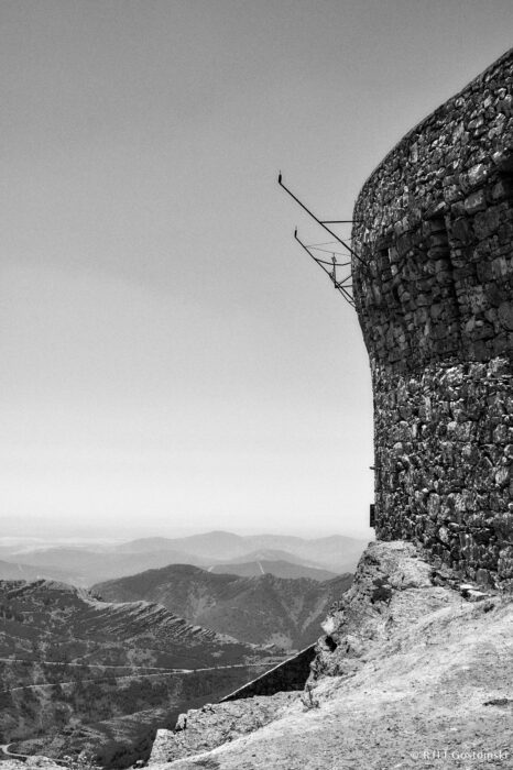 Sight markers on the Rotunda pointing at the towns and points of interest A black and white photo showing a stone rotunda wall on the right, with rugged mountain landscapes and hazy skies in the background—capturing the timeless atmosphere of Peña de Francia. Sight markers on the Rotunda point at towns and points of interest