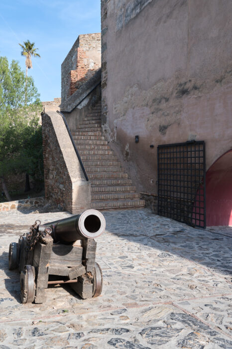 An old cannon at Castillo Gibralfaro A historic cannon on a stone courtyard sits near a staircase and an old brick wall, with greenery and a palm tree visible in the background.