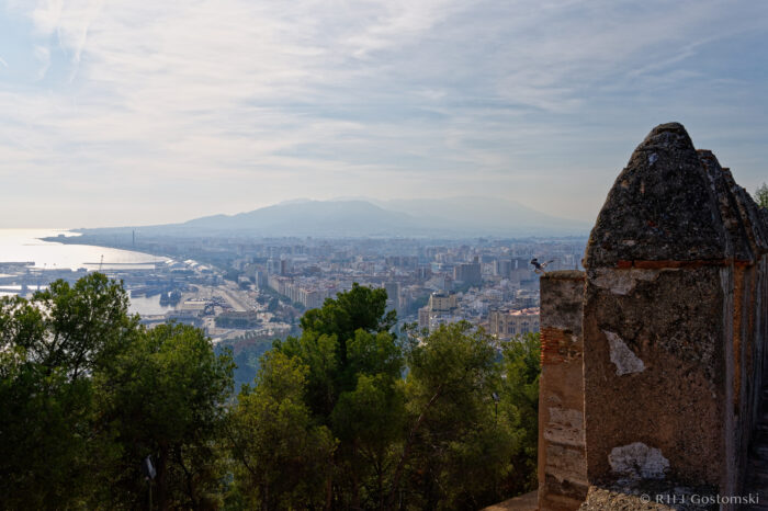 View of Malaga from Castillo Gibralfaro View of a coastal city with a harbour, tall buildings, and mountains in the background, seen from an elevated stone structure surrounded by trees under a hazy sky.