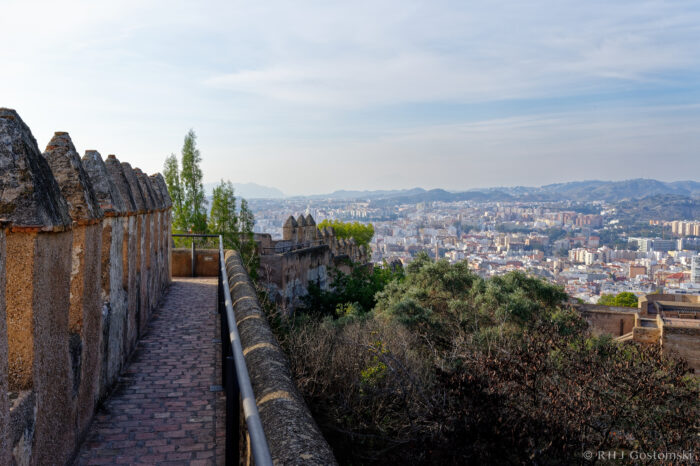 View of Malaga from Castillo Gibralfaro A stone walkway with crenellated walls overlooks a sprawling cityscape, with green trees and distant mountains under a clear blue sky.