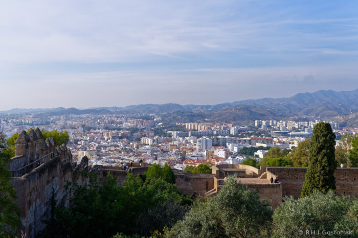 View of a city with numerous buildings and flats surrounded by hills and mountains, seen from an elevated point with ancient stone ruins and trees in the foreground under a partly cloudy sky.