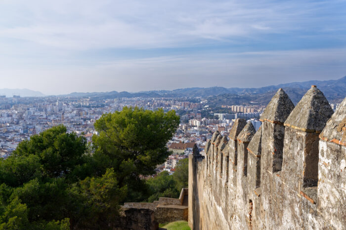 View from a stone fortress wall overlooking a city with white buildings, green trees, and distant mountains under a blue sky with wispy clouds.