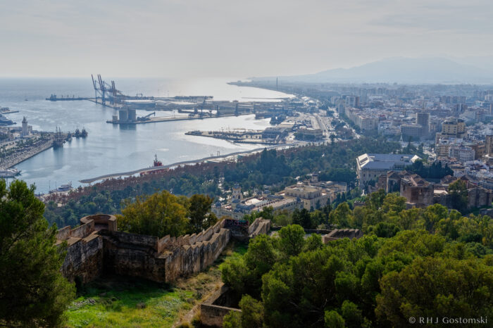 View of Malaga from Castillo Gibralfaro A panoramic view of Málaga, Spain, showing the harbour with cranes, the city skyline, the coastline, and the remains of an old fortress surrounded by greenery in the foreground.