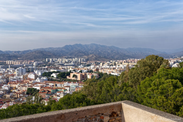 View of Malaga from Castillo Gibralfaro Cityscape with a mix of colourful buildings and green trees in the foreground, sprawling towards distant mountains under a partly cloudy blue sky.