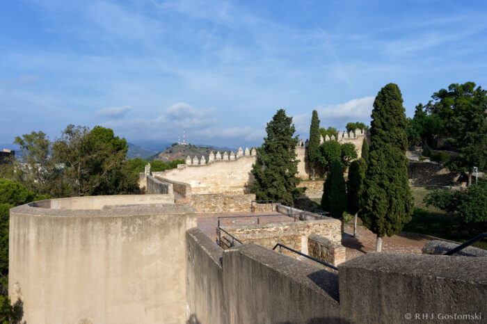 Stone ramparts and towers of a historic fortress, surrounded by green trees under a blue sky, with distant hills and communication masts visible in the background.