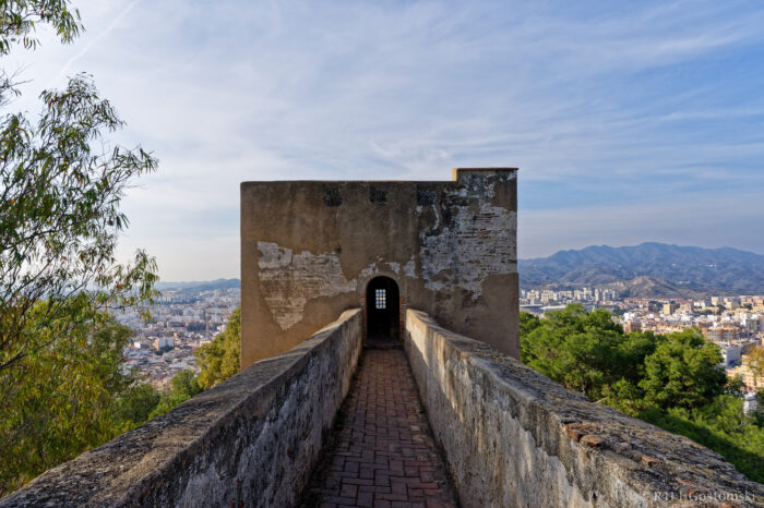 A narrow stone walkway leads to a weathered watchtower with a small window, overlooking a city surrounded by hills and trees under a partly cloudy sky.
