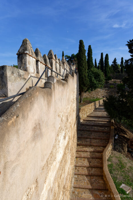 Castillo Gibralfaro rampart wall A stone stairway runs alongside an old, weathered fortress wall with pointed battlements, lined with trees under a clear blue sky.