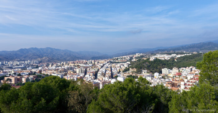View of Malaga from Castillo Gibralfaro A panoramic view of a city with clusters of white and tan buildings nestled among green hills, with mountains in the background under a partly cloudy blue sky.
