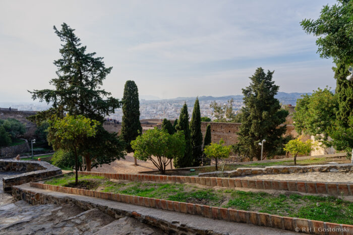 Castillo Gibralfaro A terraced stone pathway with steps, lined with trees and shrubs, leads down to a courtyard. The cityscape and distant mountains are visible under a partly cloudy sky.