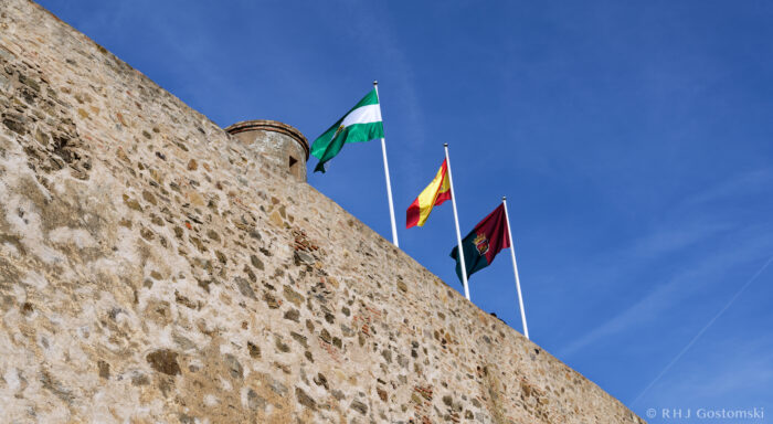 Flags flying on the rampart wall at Castillo Gibralfaro Three flags—green and white, red and yellow, and burgundy—fly atop a stone fortress wall under a clear blue sky. A cylindrical turret is visible on the left side of the wall.