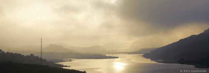 A peaceful lake winds through misty hills at sunrise, with sunlight reflecting on the water. Hills and trees appear as dark silhouettes, and a single power line crosses the scene.