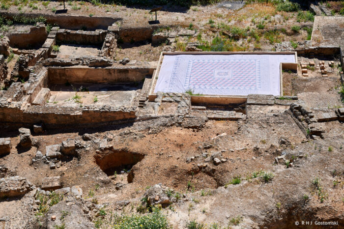 Archaeological ruins with stone walls and excavated areas, featuring a large, colourful mosaic floor in the centre, these are part of the Roman Thermal baths of Santa María, partially surrounded by grass and earth.
