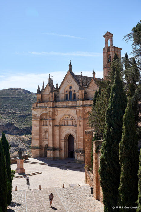 A person walks near the historic Royal Collegiate of Santa Maria, a grand stone church with pointed arches and a bell tower, surrounded by cypress trees in a sunlit, mountainous landscape.