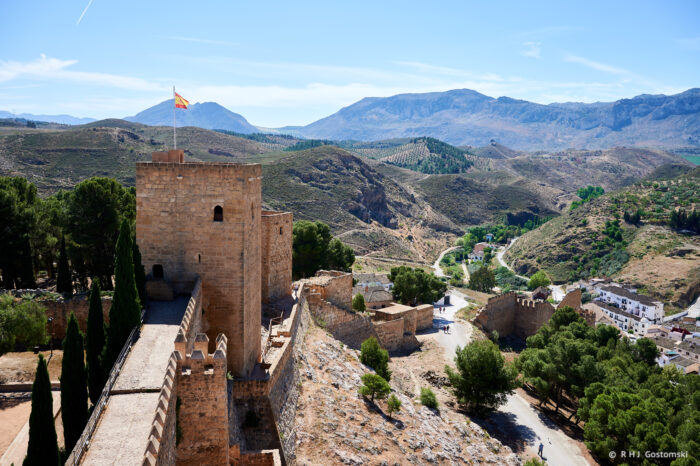 The Alcazaba in Antequera, with a Spanish flag flying on one of the watch towers, stands on a hill, surrounded by trees and mountains under a clear blue sky. A winding road and small buildings are visible in the valley below.