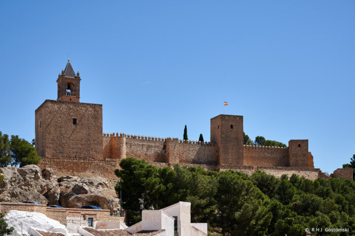 La Alcazaba in Antequera, a large stone fortress with crenellated walls and square towers, stands on a rocky hill surrounded by trees under a clear blue sky. A Spanish flag flies proudly from one of the towers.