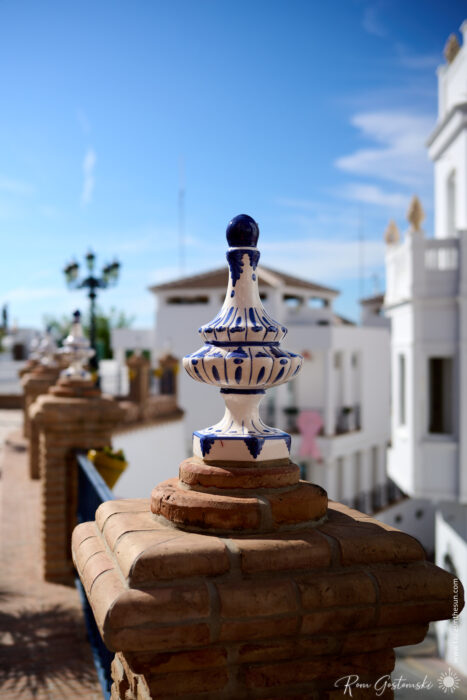 A blue and white ceramic finial sits on a brick pier, with more piers receding into the background. White buildings and a blue sky are visible, suggesting a Mediterranean or Spanish setting.