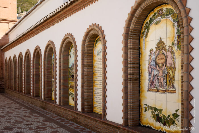 A row of arched brick-framed niches set in the white side wall of Parroquia Nuestra Se&ntilde;ora de la Asunci&oacute;n, each displaying colourful ceramic tile murals with religious or decorative scenes and text. The path is tiled, and the scene is sunlit.