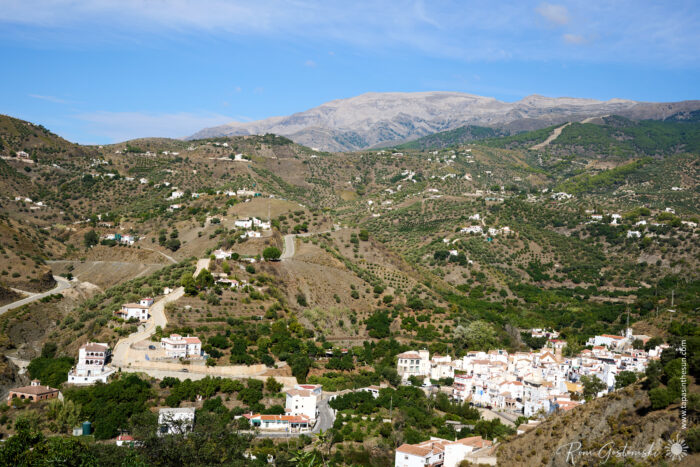 A small village with white buildings nestles among green hills and winding roads, with a mountain range in the background under a blue sky with wispy clouds.