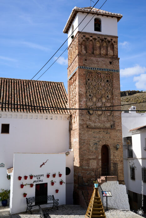 A tall, ornate brick tower&mdash;the Alminar Mud&eacute;jar de &Aacute;rchez&mdash;with Moorish details stands beside a whitewashed building adorned with red flower pots and a lizard decoration, under a blue sky with clouds and intersecting power lines.