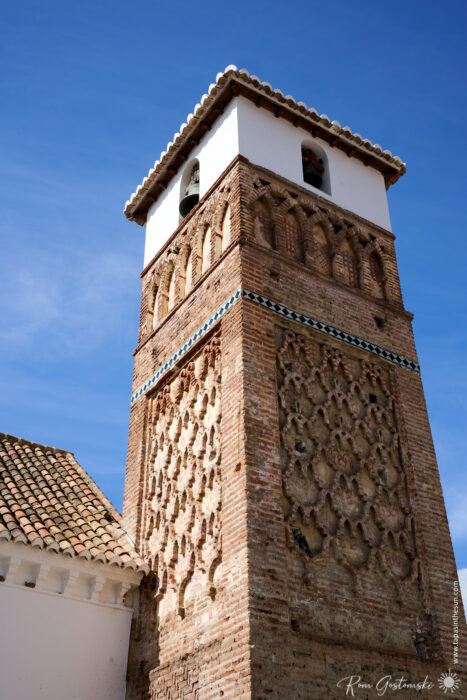 The Alminar Mud&eacute;jar de &Aacute;rchez has a tall brick bell tower with intricate geometric patterns and a white upper section, standing against a clear blue sky and attached to a building with a tiled roof.