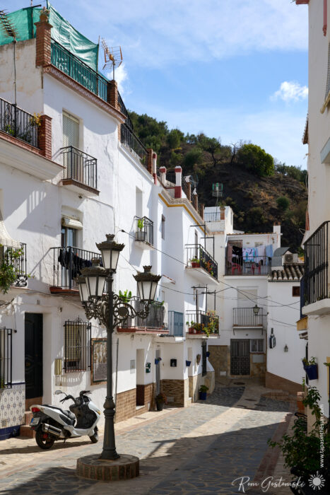 A narrow, sunlit street in &Aacute;rchez winds between whitewashed buildings with balconies and potted plants. A scooter rests nearby, while washing hangs between houses; street lamps line the way and a low hill rises in the background.
